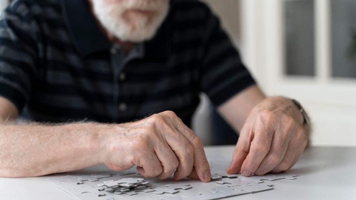 Imagem de um idoso montando um quebra-cabeça sobre uma mesa, em contexto informativo sobre Alzheimer e fatores de risco, com referência ao impacto até 2050 no Brasil.