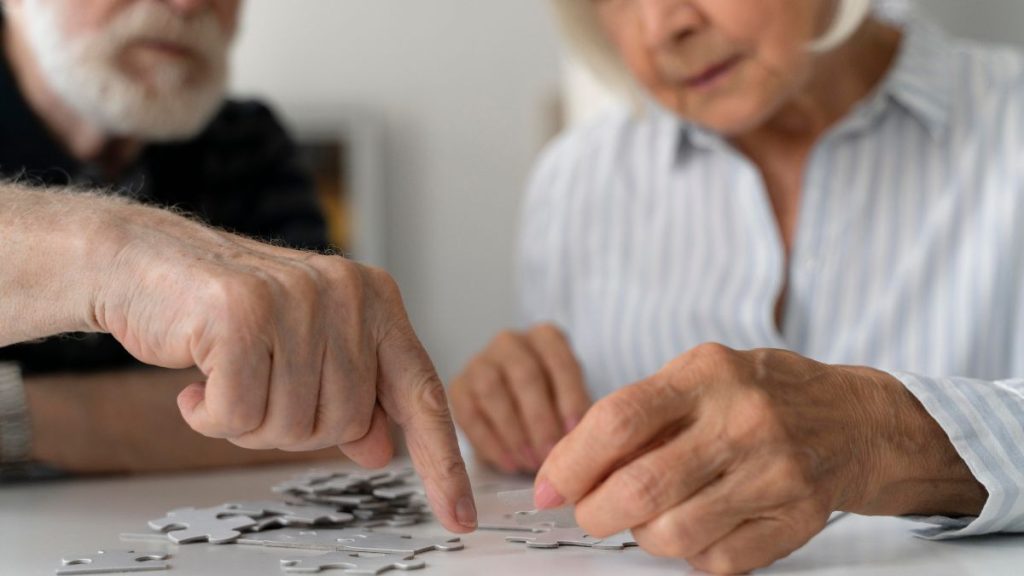 Idosos sentados à mesa, com mãos sobre peças de dominó, sugerindo atividade cognitiva e cuidado na prevenção do Alzheimer; imagem sobre fatores de risco