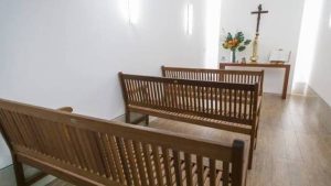 Interior de capela com bancos de madeira, piso claro e ao fundo um altar com cruz cristã e arranjos florais, ambiente religioso iluminado.