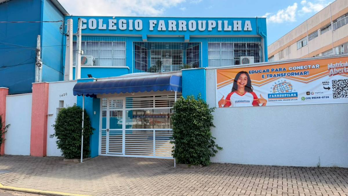 Fachada do Colégio Farroupilha, com prédio azul e faixa de entrada escrita “COLÉGIO FARROUPILHA”, além de banner educacional e muro lateral