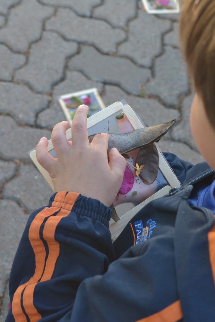 Estudante em evento cultural, segurando um material didático feito à mão sobre papel e artesanato, com fundo de pedras e apresentação ligada ao Colégio Farroupilha.