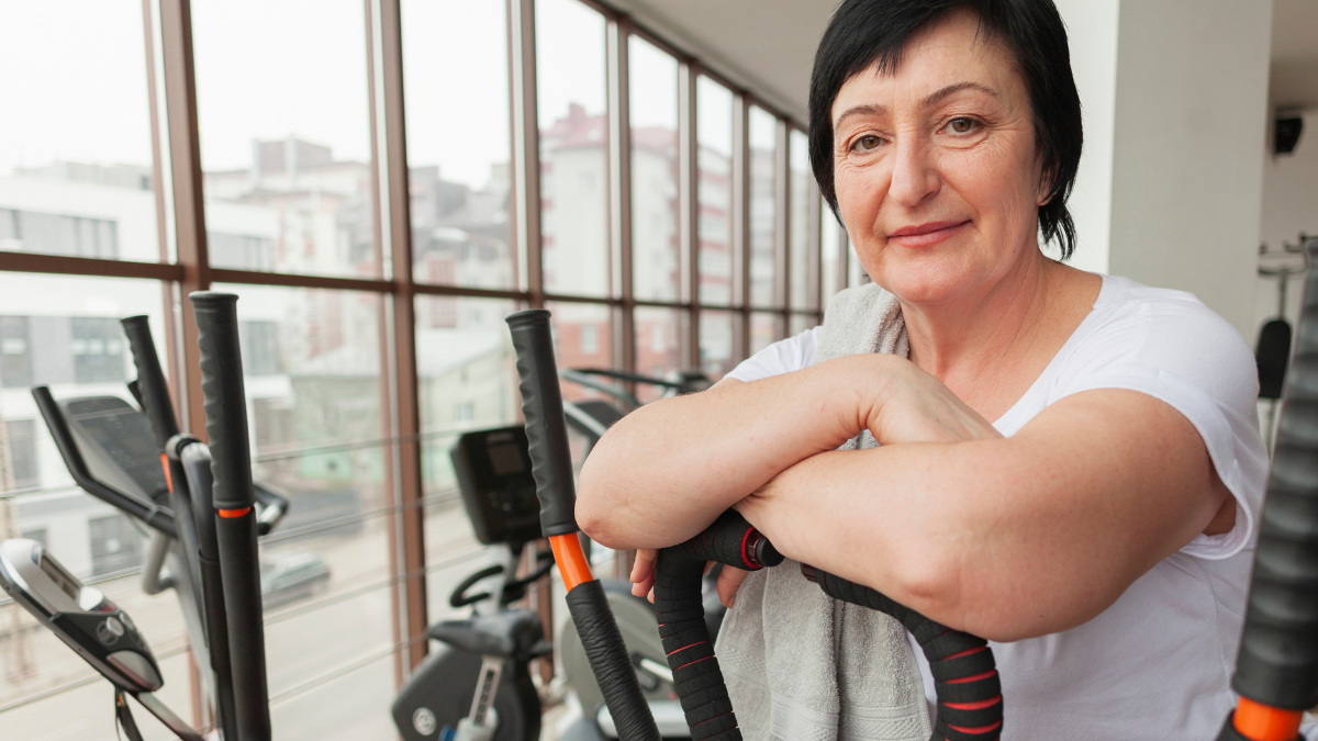 Mulher adulta em academia usa um equipamento de alongamento ou treino com alças nas mãos, em frente a janelas grandes com vista urbana ao fundo.