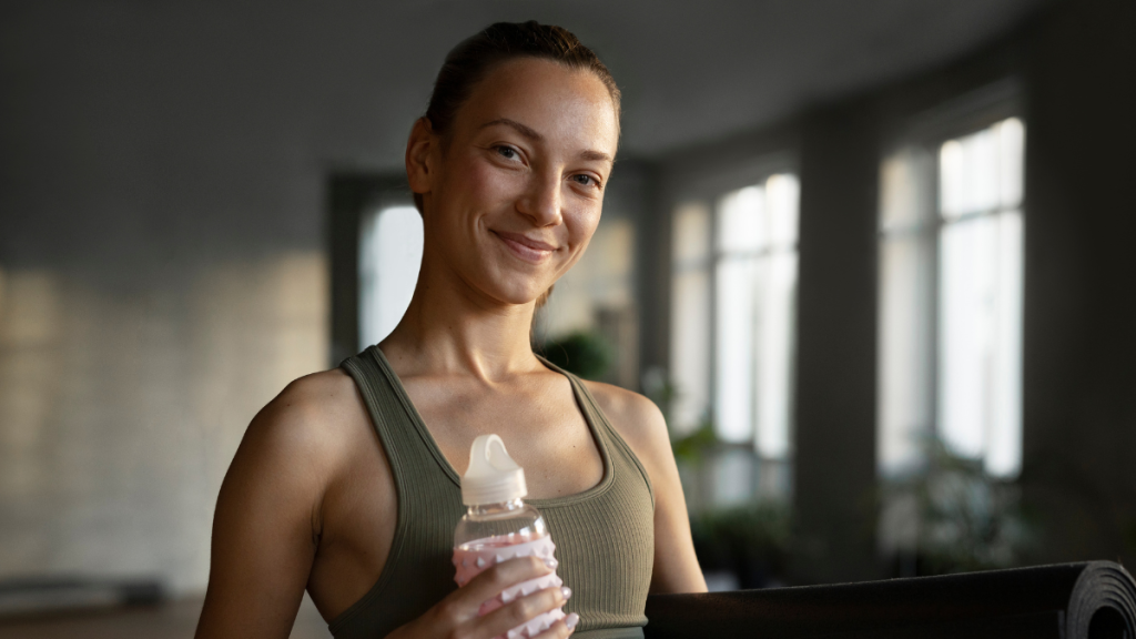 Mulher sorridente em estúdio de treino segurando um frasco de água, vestindo top esportivo, ao lado de um tapete de yoga em ambiente com luz natural.
