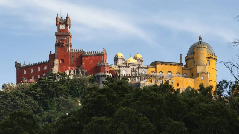 Vista panorâmica do Castelo de São Jorge em Lisboa, com torres e muralhas históricas ao lado de cúpulas amarelas sob céu claro e árvores na área frontal.