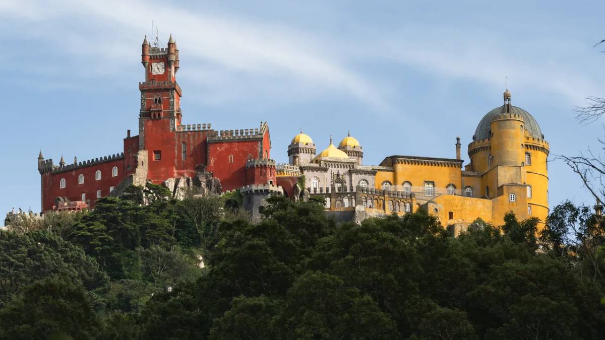 Vista panorâmica do Castelo de São Jorge em Lisboa, com torres e muralhas históricas ao lado de cúpulas amarelas sob céu claro e árvores na área frontal.