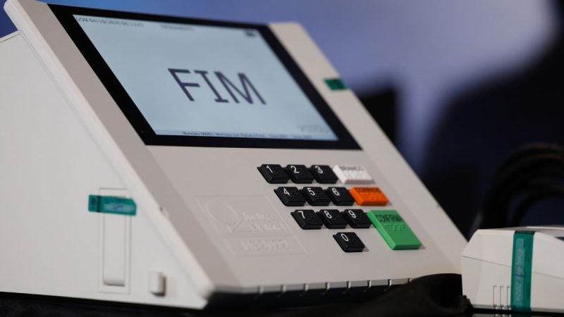 Eleições: terminal de urna eletrônica com teclas e visor ao lado de documentos, em contexto informativo sobre quem é obrigado a votar e quem tem voto facultativo