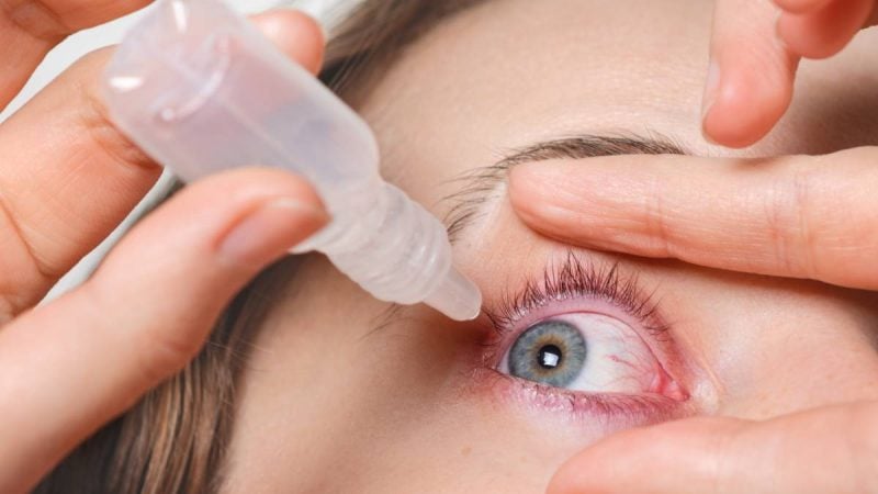 Aplicação de colírio com pipeta no olho de uma pessoa durante procedimento estético ou oftalmológico, com o olhar aberto e pálpebras sendo suavemente seguradas