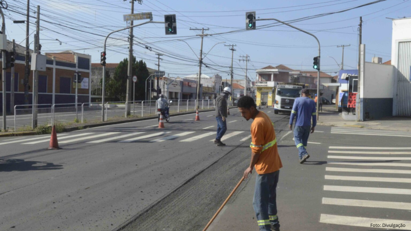 Homem varrendo e limpando uma faixa de pedestres em frente à via urbana na cidade de Piracicaba, com trabalhadores e cones de sinalização, semáforos e fiação elétrica ao fundo sob céu claro.