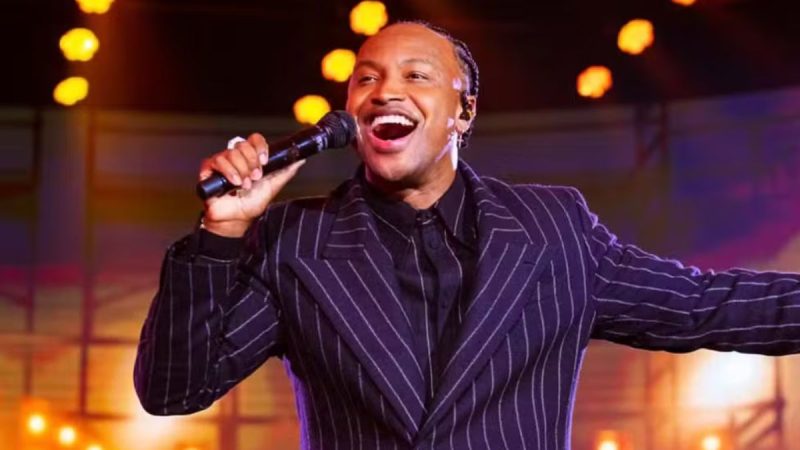 Cantor Thiaguinho em palco durante apresentação ao microfone, com expressão de alegria, vestindo terno escuro listrado e iluminação de show com luzes desfocadas ao fundo.