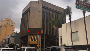 Edifício de arquitetura moderna em plano central na Avenida Francisco Glicério, em Campinas, sob céu nublado. A estrutura possui fachada de pedra e janelas verticais. Em primeiro plano, veem-se carros no trânsito urbano, palmeiras e uma placa verde de sinalização viária indicando direções para o Largo do Rosário, a Catedral e a Avenida Moraes Salles.