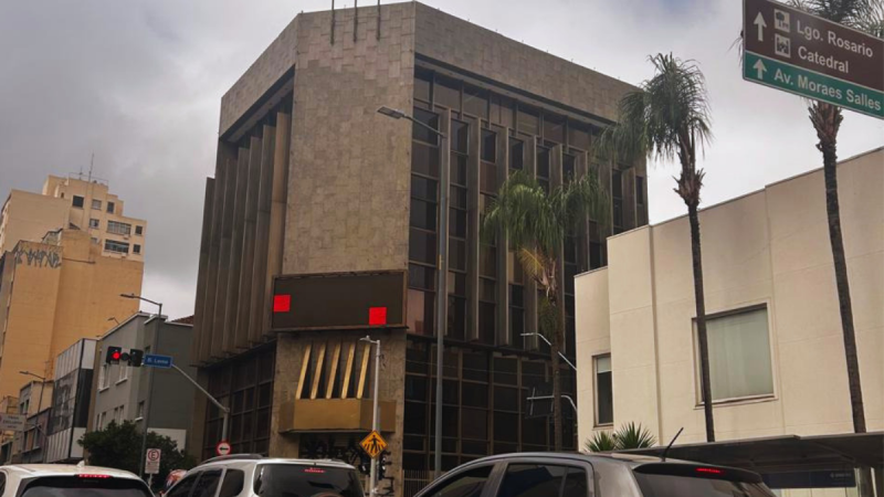 Edifício de arquitetura moderna em plano central na Avenida Francisco Glicério, em Campinas, sob céu nublado. A estrutura possui fachada de pedra e janelas verticais. Em primeiro plano, veem-se carros no trânsito urbano, palmeiras e uma placa verde de sinalização viária indicando direções para o Largo do Rosário, a Catedral e a Avenida Moraes Salles.