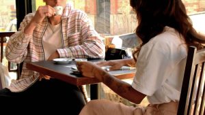 Casal em um café, com um homem e uma mulher sentados à mesa, tomando bebidas e fazendo um brinde enquanto compartilham momentos em ambiente aconchegante.