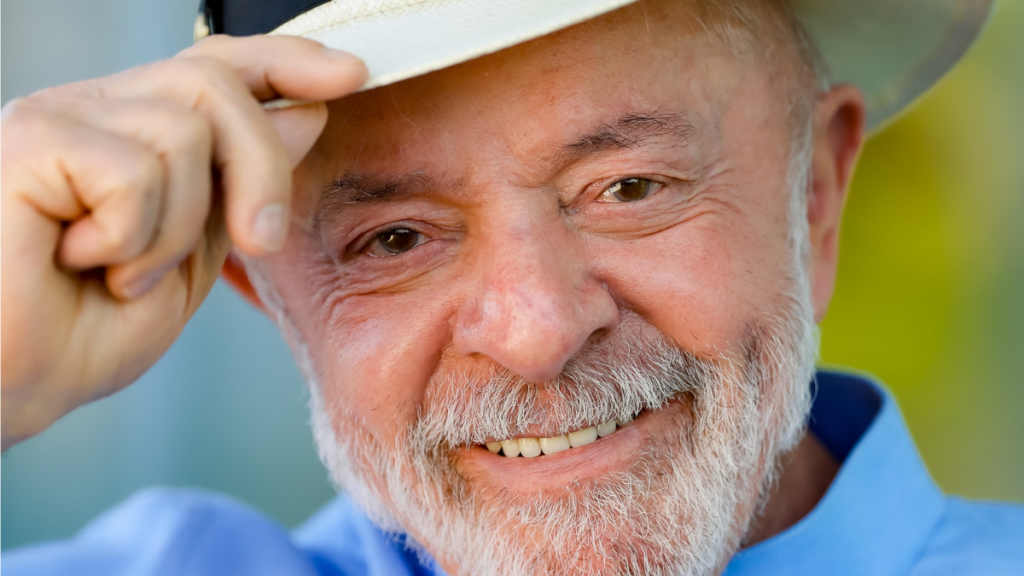 Fotografia em plano aproximado do presidente brasileiro Luiz Inácio Lula da Silva, um homem idoso de pele clara, cabelos e barba grisalhos. Ele sorri para a câmara enquanto segura com a mão direita a aba de um chapéu claro que traz na cabeça. Veste uma camisa azul clara sobre um fundo desfocado.
