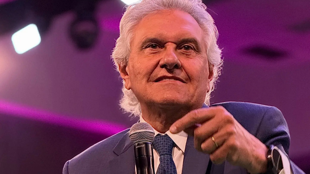 Fotografia do político brasileiro Ronaldo Caiado, com cabelo branco e fato azul, a falar ao microfone num evento iluminado em tons de roxo.