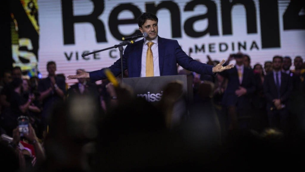 Fotografia de Renan Santos de fato azul e gravata amarela, de braços abertos num pódio a discursar num evento político, com um painel ao fundo e plateia em primeiro plano.