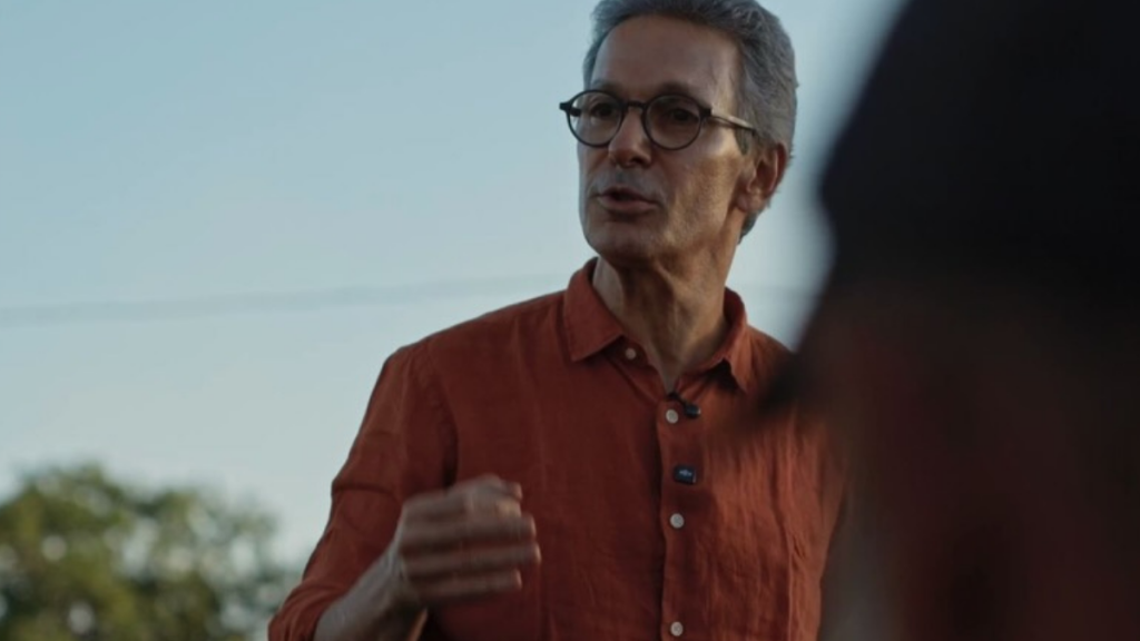 Romeu Zema, de cabelos grisalhos, óculos e camisa terracota, fala e gesticula com a mão direita em um ambiente ao ar livre com iluminação natural.