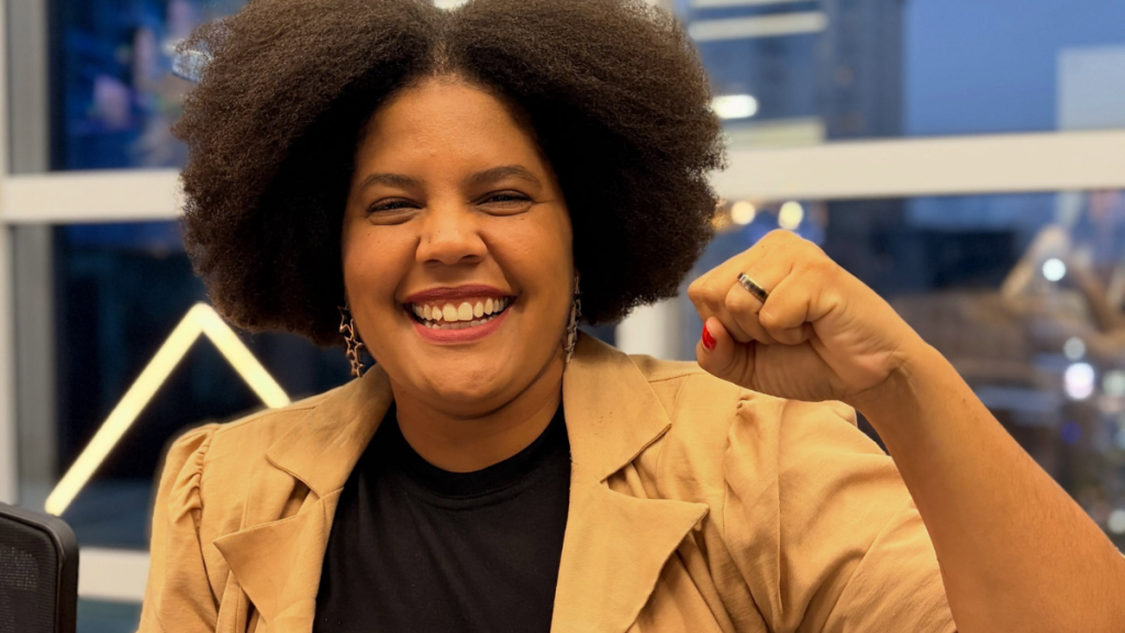 Fotografia em plano médio de Samara Martins, mulher negra sorridente com cabelo afro volumoso, a erguer o punho fechado. Veste um casaco bege sobre camisola preta e brincos em formato de estrela.