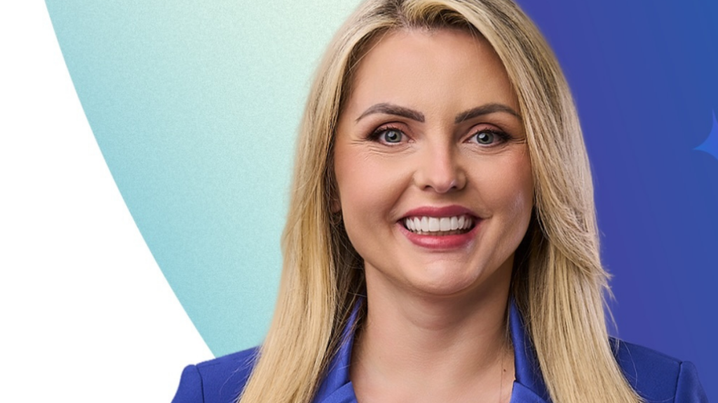 Retrato em plano aproximado de Clariana Barão, uma mulher de pele clara, olhos claros e cabelos loiros, liso e pelos ombros. Ela está a sorrir abertamente para a câmara e veste um casaco azul-escuro. O fundo apresenta tons gradientes de azul-claro, azul-escuro e branco.