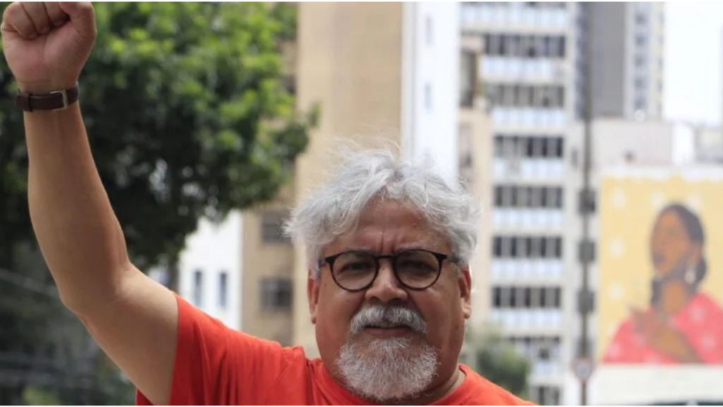 Homem de pele clara, cabelos e barba grisalhos, com óculos de armação escura, identificado como o dirigente político Edmilson Costa. Ele veste uma t-shirt vermelha com a ilustração e a frase "DEFENDER A ALEGRIA ORGANIZAR A RAIVA", e está com o braço direito erguido com o punho fechado. Ao fundo, vê-se uma paisagem urbana com edifícios e uma rua.