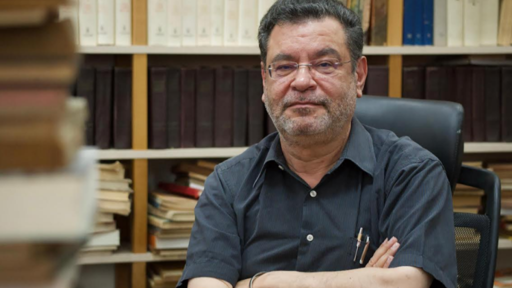 Fotografia de plano médio do político e jornalista brasileiro Rui Costa Pimenta. Ele é um homem de meia-idade, com cabelos curtos e escuros, barba grisalha e óculos de grau. Está sentado numa cadeira de escritório preta com os braços cruzados, vestindo uma camisa preta de manga curta com canetas no bolso. Ao fundo e em primeiro plano, veem-se estantes cheias de livros, criando um ambiente de biblioteca ou escritório de estudo.
