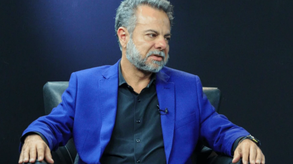 Homem maduro de pele clara, identificado como Wilson Grassi Júnior — médico-veterinário, empresário e político —, com cabelos e barba grisalhos, vestindo um blazer azul-marinho sobre uma camisa preta. Encontra-se sentado numa poltrona de couro preta, num cenário de estúdio escuro com iluminação focada.