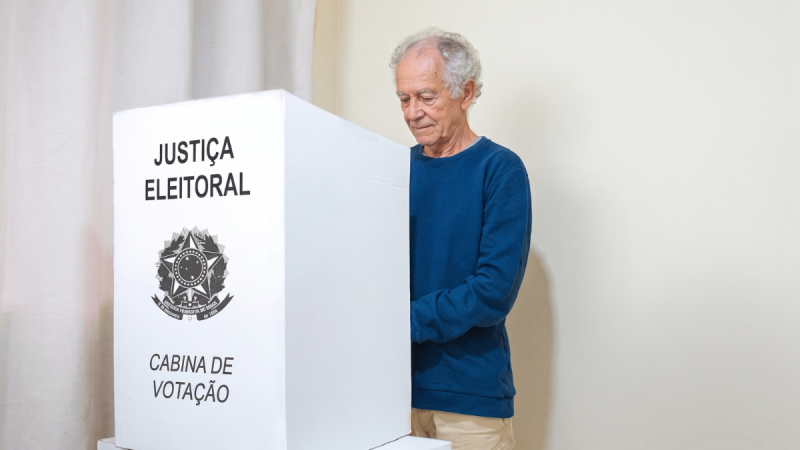 Homem idoso de cerca de 70 anos, com cabelos brancos e cacheados, de pé atrás de uma cabina de votação branca da Justiça Eleitoral brasileira. Ele veste blusa azul de manga comprida e calça bege, enquanto vota. O fundo é uma parede neutra clara.