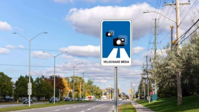 Placa de sinalização de velocidade média em uma avenida, com postes de iluminação e carros ao fundo sob céu parcialmente nublado.