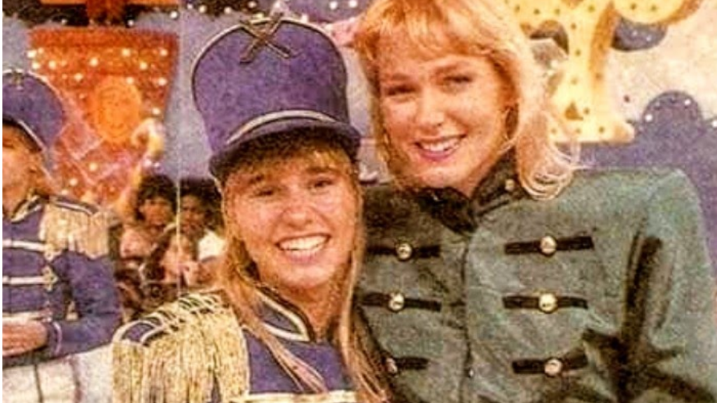 Fotografia de arquivo do programa infantil Xou da Xuxa. Em primeiro plano, a apresentadora Xuxa Meneghel surge sorridente ao lado de Andréa Sorvetão vestida com o fato característico de soldado de infantaria, composto por casaco azul com detalhes dourados e barretina. Ao fundo, observa-se parte do cenário colorido do programa e crianças na plateia.