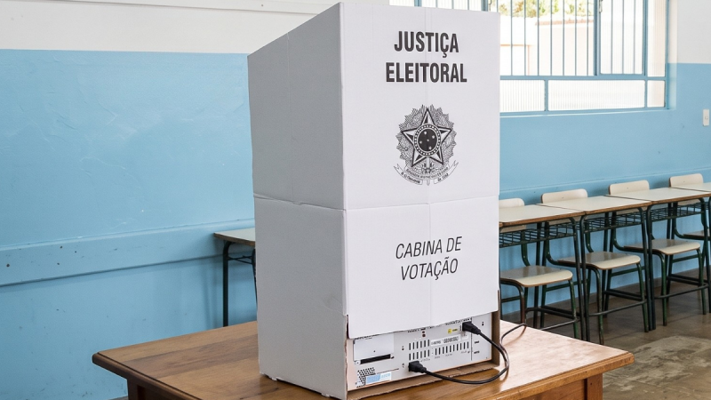 Uma cabine de votação de papelão branco da Justiça Eleitoral do Brasil, colocada sobre uma mesa de madeira no centro de uma sala de aula. Na parte superior da cabina, destaca-se o brasão da República Federativa do Brasil ladeado pelas palavras "JUSTIÇA ELEITORAL", e na parte inferior lê-se "CABINA DE VOTAÇÃO". Ao fundo, veem-se paredes pintadas de azul e branco e várias carteiras escolares alinhadas.