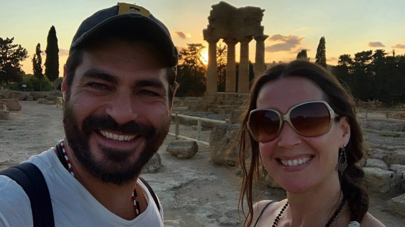Selfie do ator Thiago Lacerda e da atriz Vanessa Lóes, sorridentes ao pôr do sol nas ruínas de um templo grego antigo (Agrigento, Sicília). Thiago usa boné e camiseta branca, e Vanessa usa óculos de sol grandes.