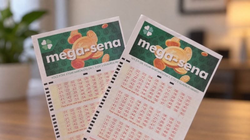 Bilhetes de loteria com o texto Mega-Sena, com cartões verdes e numeração em tabelas coloridas, apoiados em uma mesa em ambiente interno