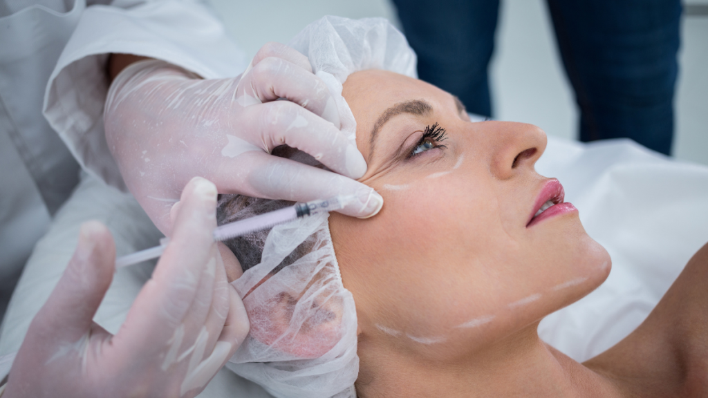 Profissional realizando aplicação de procedimento estético no rosto, com máscara e luvas descartáveis, usando seringa e gaze em paciente deitada em ambiente clínico.