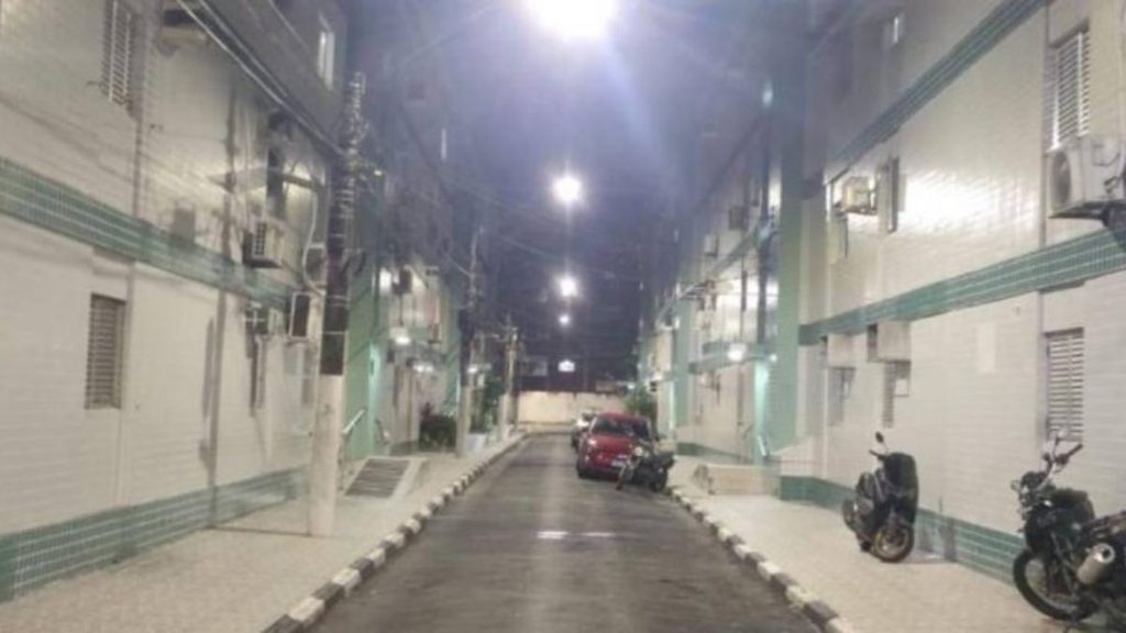 Corredor interno de prédio em Santos à noite, com motocicletas estacionadas, carros ao fundo e iluminação forte, ilustrando a notícia sobre Suspeito de matar homem no BNH em Santos e prisão temporária