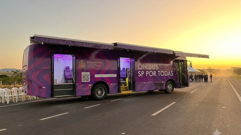 Ônibus moderno com iluminação roxa e adesivo “ÔNIBUS SP POR TODAS”, portas abertas em rodovia ao pôr do sol, com pessoas aguardando na pista.