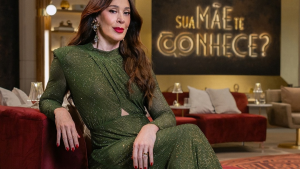 Fotografia da atriz e apresentadora Cláudia Raia posicionada no centro da imagem, sentada no braço de um sofá vermelho num estúdio de televisão elegante. Usa um vestido de mangas compridas verde-escuro brilhante com pormenores em relevo e brincos grandes prateados. Ao fundo, sob uma parede escura, destaca-se a frase em néon dourado brilhante: Sua Mãe Te Conhece?