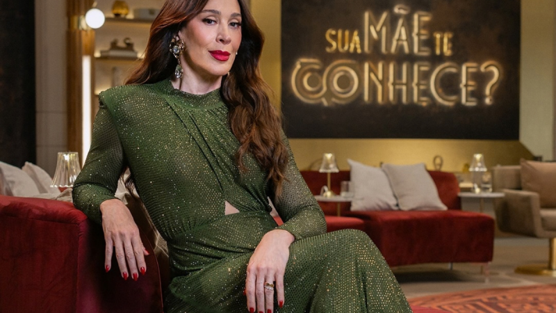 Fotografia da atriz e apresentadora Cláudia Raia posicionada no centro da imagem, sentada no braço de um sofá vermelho num estúdio de televisão elegante. Usa um vestido de mangas compridas verde-escuro brilhante com pormenores em relevo e brincos grandes prateados. Ao fundo, sob uma parede escura, destaca-se a frase em néon dourado brilhante: Sua Mãe Te Conhece?