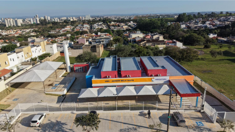 Fotografia aérea da Unidade Básica de Saúde (UBS) do Jardim Bela Vista, em Indaiatuba. O edifício moderno de piso único possui fachadas em tons de vermelho, laranja e azul, com a inscrição "UBS - JARDIM BELA VISTA" numa faixa amarela. À frente da entrada, destaca-se uma área coberta por várias tendas brancas piramidais e um estacionamento com vagas delimitadas. Ao fundo e em redor, observa-se a paisagem urbana do bairro, composta por casas, edifícios e áreas verdes.