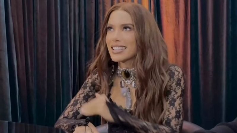 Anitta aparece em entrevista, com expressão confiante e em um ambiente de estúdio com cortinas ao fundo, falando ao lado de uma mesa.