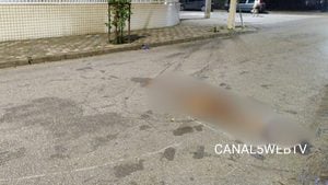 Acidente em Santos: área de rua com marca de óleo e um corpo coberto após colisão envolvendo moto, bicicleta e pedestre, com cena de atendimento próximo a calçada e muro