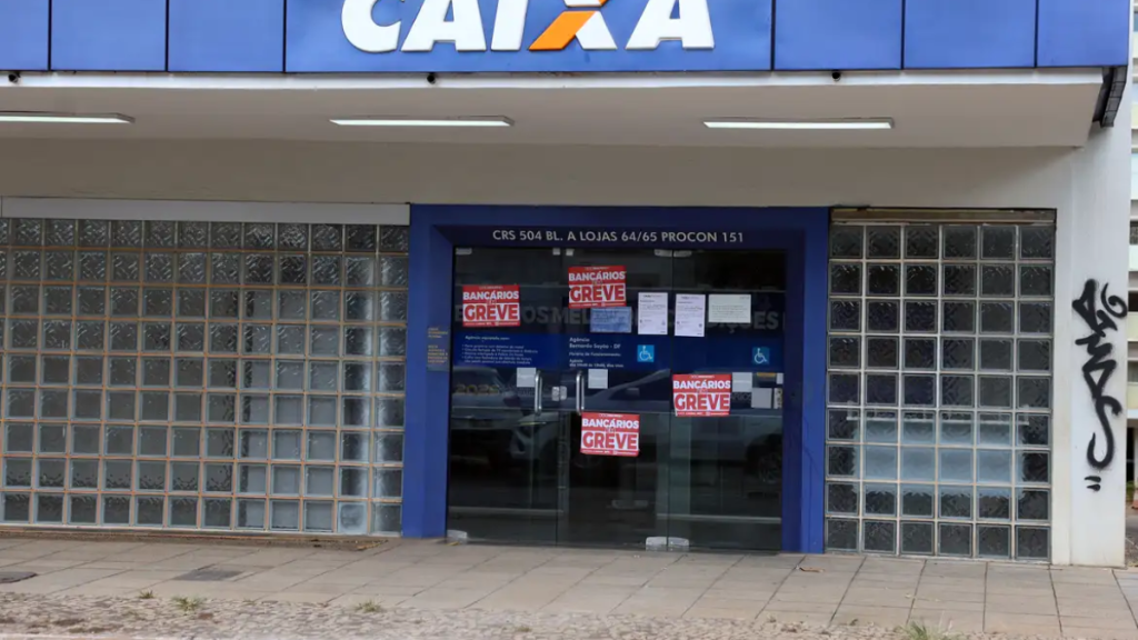 Fachada de loja Caixa com cartazes de “Bancários em greve” no vidro da entrada, placas e avisos internos visíveis, em prédio comercial.