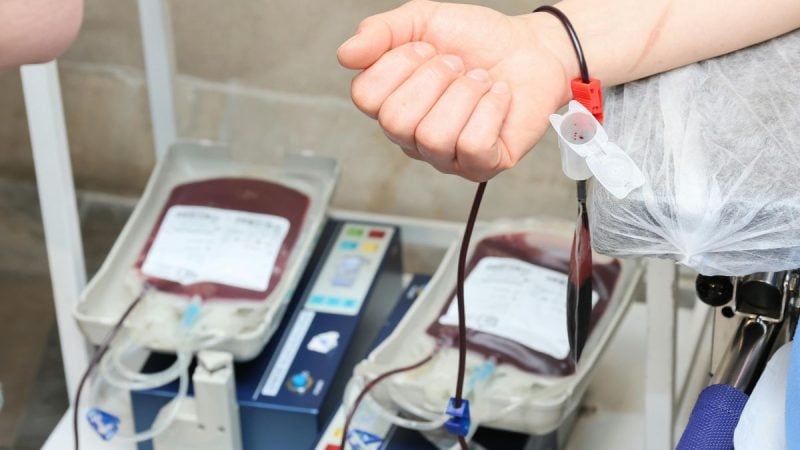 Mão com equipo de transfusão de sangue em ambiente hospitalar, com bolsas e equipamentos ao fundo, representando regras flexibilizadas sobre doação e transfusão de sangue por Testemunhas de Jeová.