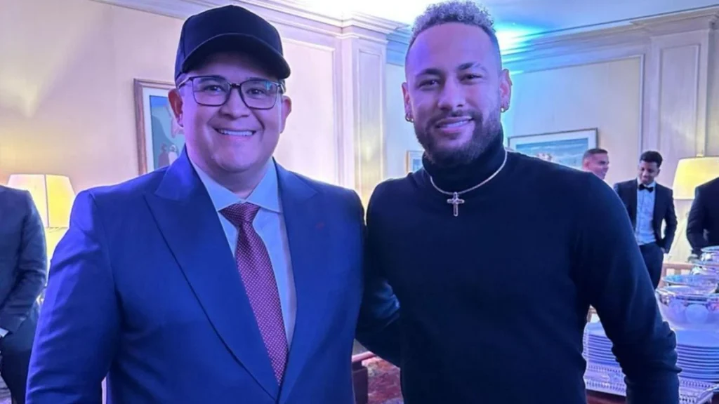 Bruno Avelar, de terno, e Neymar, de moletom escuro, posando sorridentes ao lado de convidados em ambiente interno.