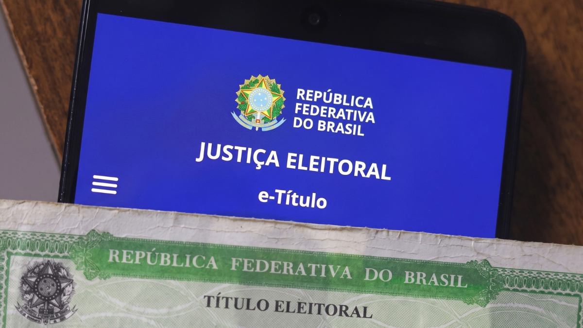 Smartphone a apresentar no ecrã a aplicação digital e-Título da Justiça Eleitoral brasileira com fundo azul, posicionado sobre uma versão impressa em papel do Título Eleitoral em tom verde.