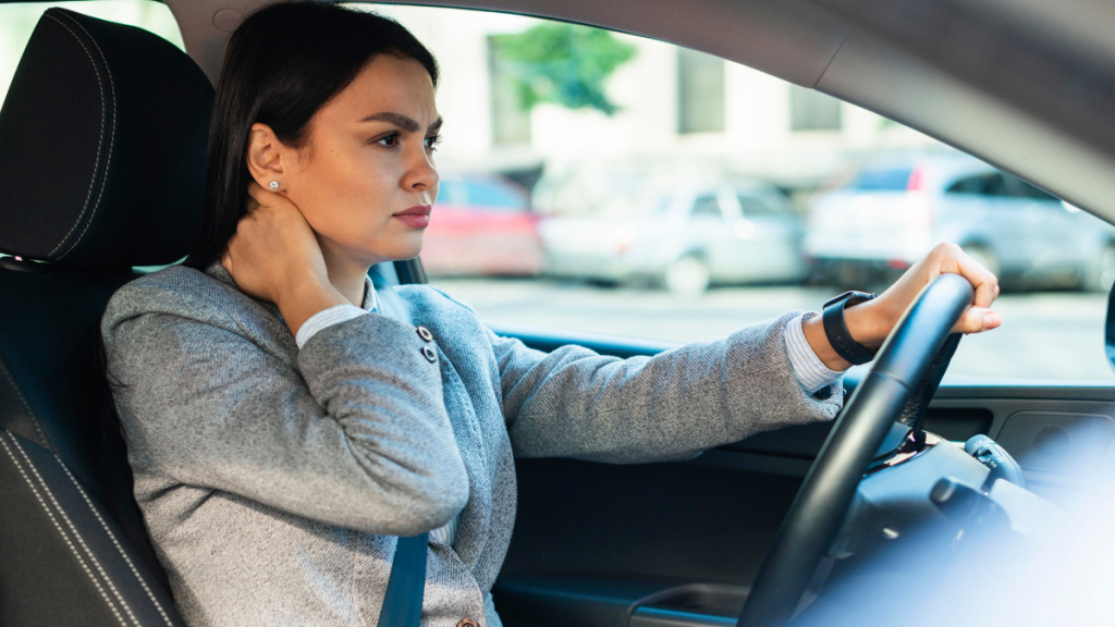 Mulher dirigindo um carro, segurando o volante com as duas mãos, em um ambiente urbano ao fundo. Foto em perspectiva do banco do motorista.