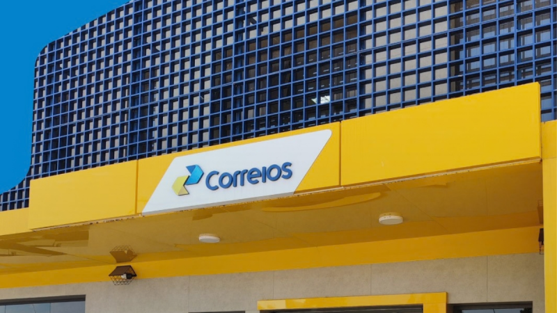 Fachada amarela de uma agência dos Correios com a logótipo oficial em azul e amarelo sobre uma placa branca. O edifício apresenta uma estrutura metálica em grelha azul na parte superior, sob um céu azul limpo.