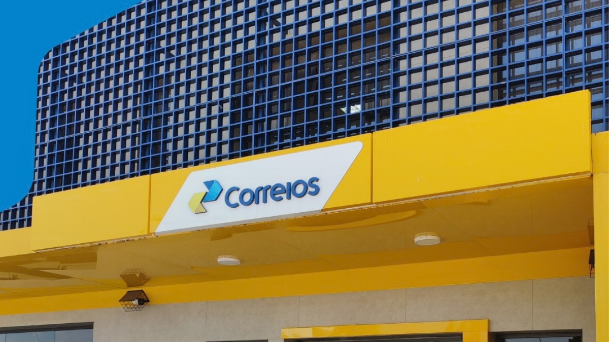 Fachada amarela de uma agência dos Correios com a logótipo oficial em azul e amarelo sobre uma placa branca. O edifício apresenta uma estrutura metálica em grelha azul na parte superior, sob um céu azul limpo.
