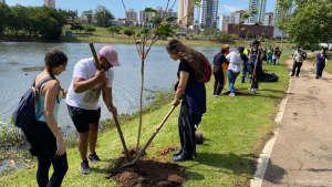 Um grupo de pessoas participa na plantação de árvores na cidade de Indaiatuba, numa área verde ao lado de um lago num parque urbano. Em primeiro plano, uma mulher de blusa clara e mochila, um homem de t-shirt branca e boné rosa com uma pá, e outra mulher de vestuário escuro trabalham em conjunto para plantar uma muda de árvore na terra. Ao fundo, veem-se outras pessoas a realizar a mesma atividade ao longo do caminho, com o lago à esquerda e edifícios da cidade no horizonte.
