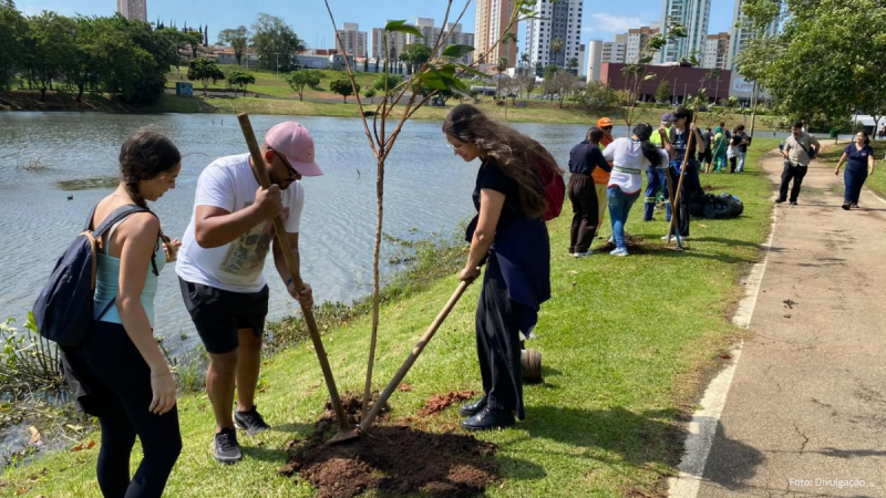 Um grupo de pessoas participa na plantação de árvores na cidade de Indaiatuba, numa área verde ao lado de um lago num parque urbano. Em primeiro plano, uma mulher de blusa clara e mochila, um homem de t-shirt branca e boné rosa com uma pá, e outra mulher de vestuário escuro trabalham em conjunto para plantar uma muda de árvore na terra. Ao fundo, veem-se outras pessoas a realizar a mesma atividade ao longo do caminho, com o lago à esquerda e edifícios da cidade no horizonte.