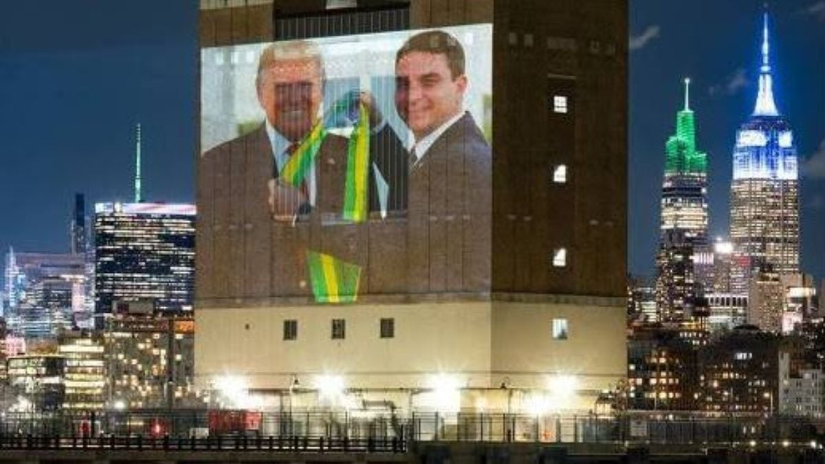 Prédio corporativo durante a noite com telão exibindo dois homens em transmissão ou evento, enquanto ao fundo a skyline iluminada de uma cidade com edifícios e torres se destaca.