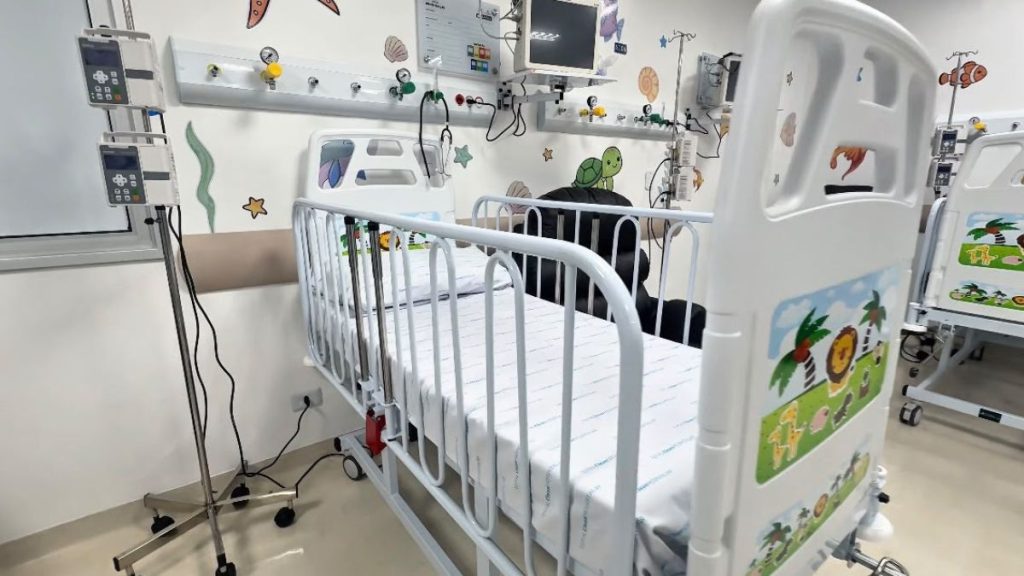 Quarto hospitalar infantil com leito pediátrico branco, paredes claras com adesivos coloridos de animais e equipamentos médicos ao fundo, em ambiente de cuidados